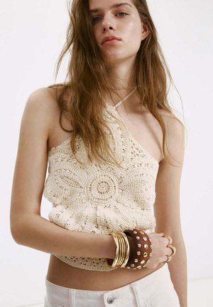 Joven mujer con un top halter de crochet color crema, pantalones blancos, pulseras doradas y una pulsera ancha marrón con tachuelas en su muñeca izquierda.