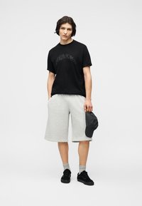 Νέος άνδρας στέκεται φορώντας μαύρο γραφικό μπλουζάκι, ανοιχτό γκρι σορτς μέχρι το γόνατο, γκρι κάλτσες, μαύρα αθλητικά παπούτσια, κρατώντας μαύρο καπέλο στο δεξί χέρι.