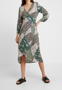 Vestido de patrón floral en tonos variados de verde, azul marino y blanco; con escote en V, mangas largas y una abertura lateral. Llevado con sandalias negras con tachuelas.
