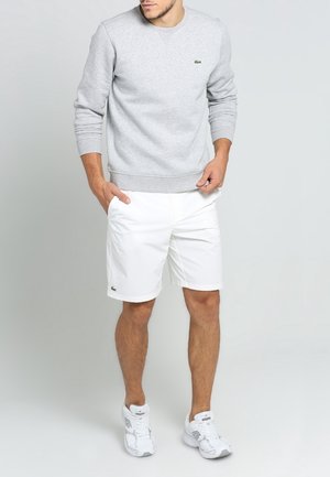 Man wearing light grey sweatshirt, white shorts, and white sneakers standing with one hand in pocket and the other adjusting sweatshirt hem.