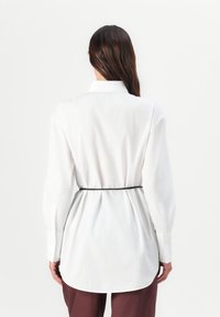 White long-sleeve shirt with a pointed collar, belted at the waist with a black strap, featuring a curved hem and smooth fabric.