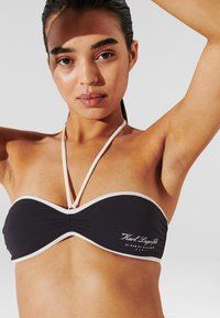 Crni bandeau bikini top s bijelim oblogom, izrađen od elastičnog materijala i s nabiranjem na sredini. Izvezeni logo sa strane.