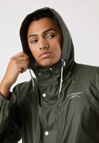 Olijfgroene waterdichte jas met capuchon, zilveren drukknopen en treklint. Voorzien van elastische manchetten en logodetails op de borst.