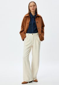 Chaqueta de ante marrón, camisa de mezclilla, pantalones de pierna ancha texturizados en crema y botas de ante marrón hasta el tobillo. Botones visibles en la chaqueta y los pantalones.
