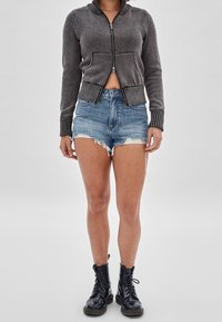 Szary rozpinany sweter z dzianiny z prążkowanym kołnierzem, zestawiony z postrzępionymi niebieskimi szortami jeansowymi i czarnymi butami na sznurowadła.