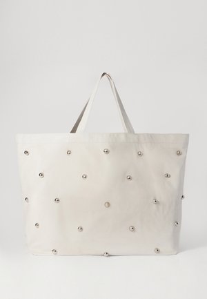 Beżowa torba na ramię z równomiernie rozmieszczonymi srebrnymi ćwiekami na powierzchni. Posiada dwie górne rączki oraz prosty, niepodszewkowany design wnętrza.