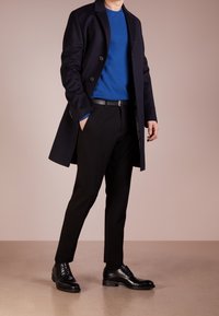 Black wool coat over a blue sweater, paired with tailored black trousers and black lace-up shoes. Smooth textures and fitted silhouette.