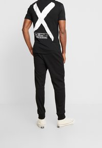 Camiseta negra con un gran "X" blanca y diseño de texto en la parte posterior. Combinada con pantalones jogger negros y zapatillas de deporte de color claro.