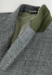 Grijze blazer met een tekstuurgedetailleerd ruitpatroon, voorzien van een groene vilt kraag en een gladde binnenvoering. Gedetailleerde stiksels en een getailleerde pasvorm.