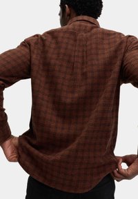 Camisa a cuadros marrón con un tejido texturizado, cuello abotonado y mangas largas. La tela presenta un sutil patrón de fondo de perro.