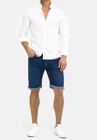 Camisa de lino blanca con botones y pantalones cortos de denim con los puños enrollados. Las zapatillas son blancas. Look general casual con un contraste de colores claros y oscuros.