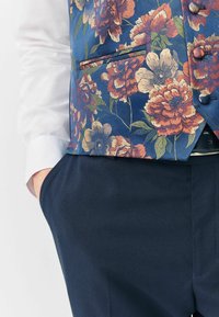 Blumenveste aus blauem Stoff mit orangefarbener und cremefarbener Blüten. Verfügt über eine einzelne Tasche und Knopfdetails, kombiniert mit dunkelblauen Hosen.