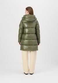 Olijfgroene pufferjack met een glanzende, soepele textuur, oversized capuchon, gestikt patroon en knielange snit, gecombineerd met crèmekleurige broeken.