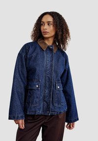 Jeans jakna u tamnoplavoj boji s smešnim baršunastim ovratnikom, dva prednja džepa i vidljivim šavovima. Labav kroj s širokim rukavima.