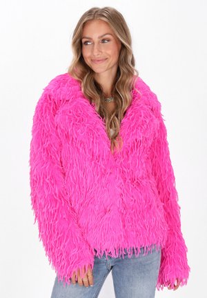 Veste en fausse fourrure rose avec une texture shaggy, un large col, des manches longues et des détails en frange au niveau de l'ourlet, associée à un jean bleu clair.