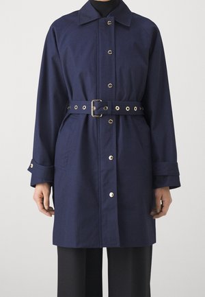 Marineblå bæltefrakke med sølvknapper, lange ærmer og bred krave båret over sorte bukser og en sort top.