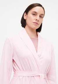 Lyse rosa striped morgenkåpe med sjalkrage, belte og myk tekstur, båret over en lyse rosa topp. Har diskrete logo-detaljer.