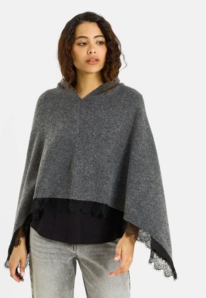Mujer con cabello rizado que lleva un poncho gris con capucha con ribete de encaje negro sobre una camisa negra y jeans gris claro, de pie frente a un fondo blanco.