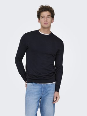 Mann med krøllete hår står mot hvit bakgrunn iført en svart langermet skjorte over en hvit t-skjorte og lyseblå jeans.