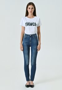 T-shirt bianca con testo nero "cose belle", abbinata a jeans skinny blu a vita alta. Il modello indossa ballerine nere.