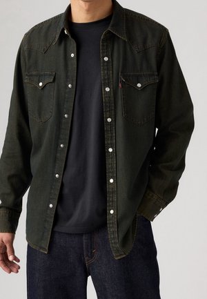 Homme portant une chemise en jean boutonnée vert foncé sur un t-shirt noir et un jean bleu foncé, debout avec une main dans la poche sur un fond uni.
