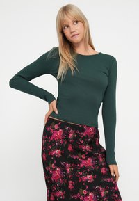 Ribbede, langærmede top i dyb grøn, parret med en sort blomsterkjole med livlige pink blomster og grønne accenter.