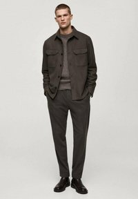 Dark, textured jacket with two chest pockets, worn over a light grey knit sweater and slim-fitting, dark trousers, paired with black boots.