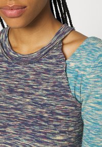 Kvinna i lager av ribbstickade toppar i flerfärgat lila och blått med ett utskärningsdetalj på axeln.