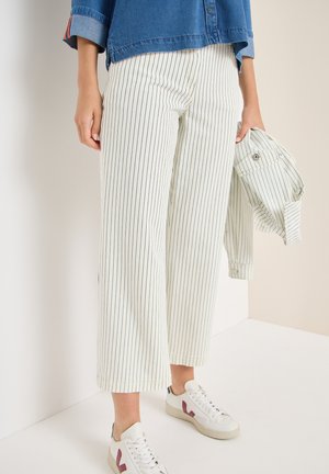Personne portant un pantalon blanc à fines rayures noires verticales, des baskets blanches avec un détail rose, tenant une veste assortie rayée, chemise bleue.