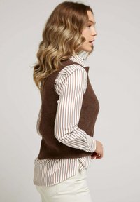 Gilet en laine marron avec une texture douce, superposé sur une chemise blanche à rayures verticales marron. Design simple et ajusté sans détails supplémentaires.