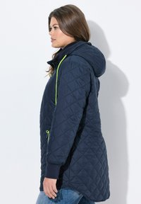 Giacca trapuntata blu navy con cappuccio, dotata di vivaci dettagli verdi lungo la zip e le cuciture, e polsini a coste. Visualizzazione laterale.
