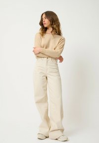 Pullover beige in maglia abbinato a pantaloni a vita alta e a gamba larga color crema. Le scarpe sono di colore chiaro con una suola testurizzata. Design semplice e minimalista.