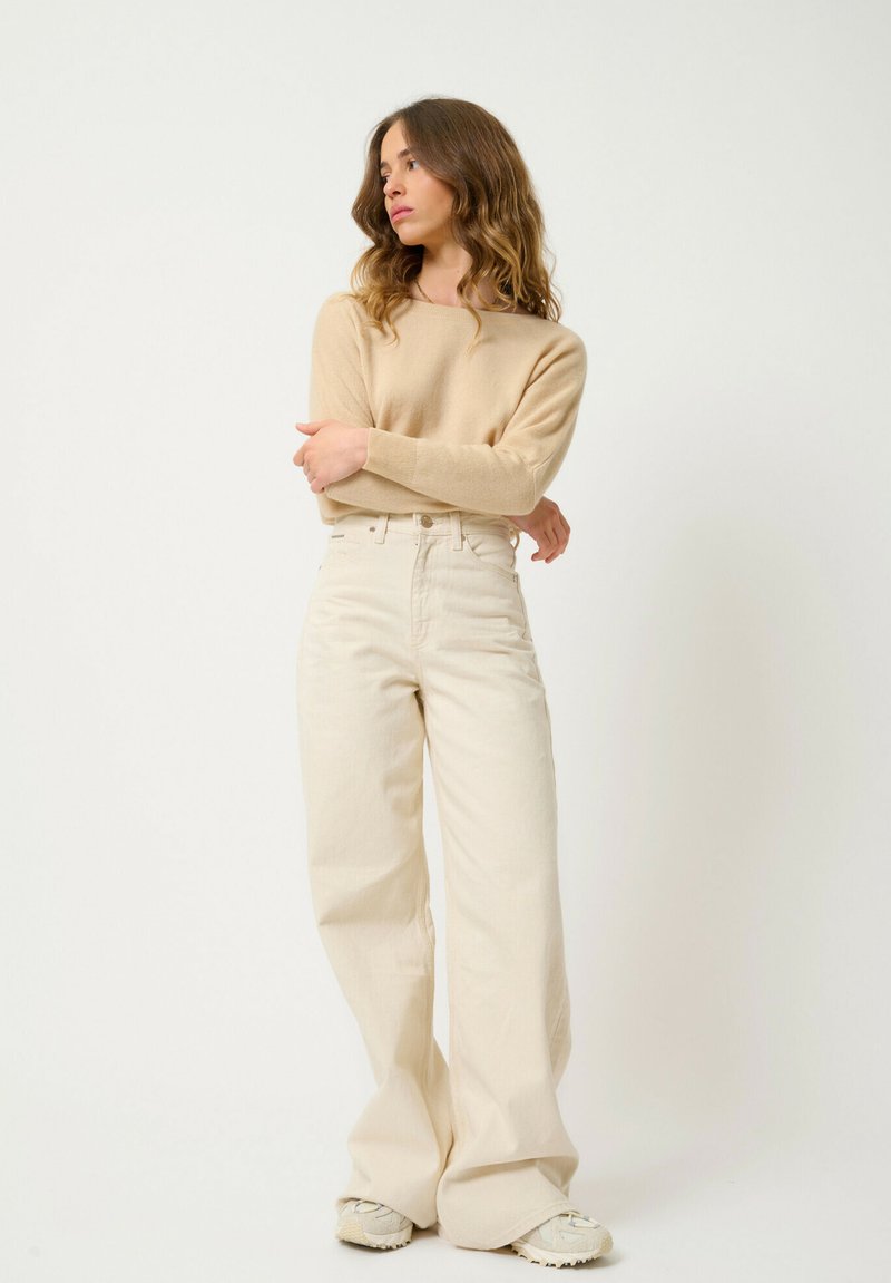 Pullover beige in maglia abbinato a pantaloni a vita alta e a gamba larga color crema. Le scarpe sono di colore chiaro con una suola testurizzata. Design semplice e minimalista.