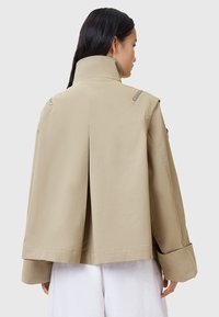 Giacca beige, corta, con colletto alto, maniche ampie e spacco posteriore; presenta zip in metallo sulle spalle e una texture liscia.