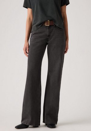 Person wearing dark gray wide-leg jeans with a brown belt and black shoes, standing against a plain light background.