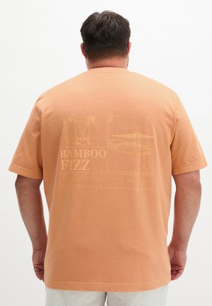 Mężczyzna w brzoskwiniowej koszulce z delikatnym nadrukiem rośliny w szklance i napisem „Bamboo Fizz” na plecach, stojący na jednolitym tle.