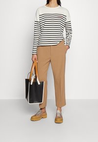 Randig långärmad tröja i vitt och svart, tillsammans med beige byxor och kraftiga beige skor. Håller en svart tote-väska med beige handtag.