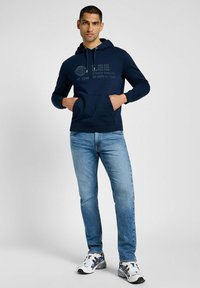 Granatowy hoodie z kieszenią kangurką i grafika w ciemnoszarym kolorze. Zestawiony z jasnoniebieskimi dżinsami i białymi trampkami z czarnymi akcentami.