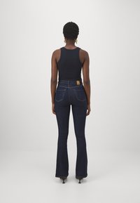 Jeans acampanados de color azul oscuro con cintura alta, costuras perfiladas, dos bolsillos traseros y una etiqueta marrón en la cinturilla. Combinados con una camiseta sin mangas negra.