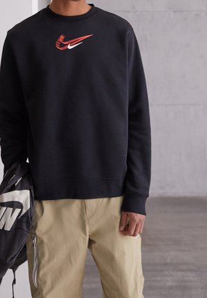 Osoba nosząca czarną bluzę z czerwonymi i białymi logo Nike, beżowe spodnie, trzymająca czarną kurtkę Nike z białym logo.