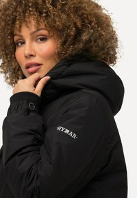 Doudoune noire avec capuche, arborant un logo sur la manche et une texture de tissu lisse et brillante. Gros plan sur la zone du col.