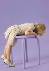 Lila rund pall med cylindriska ben. Ett barn i en mönstrad krämfärgad klänning lutar med armarna i kors på sätet. Mjuka texturer synliga.