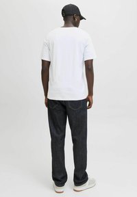 Hvid kortærmet t-shirt, mørke denim jeans og grå sneakers. Sorte cap på hovedet bagfra, med en metallisk ørering synlig.
