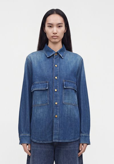 Jeune personne aux longs cheveux foncés portant une chemise en denim bleue avec boutons et deux poches à rabat sur le devant, debout devant un fond uni.