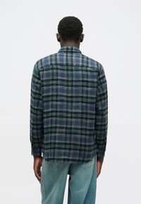 Ένα καρό φούτερ σε σκούρο πράσινο, μπλε και γκρι, με κλασικό γιακά και μακριά μανίκια, διαθέτοντας και γιακά στους ώμους και κουμπιά στους καρπούς.