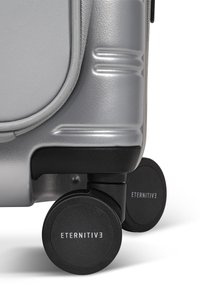 Silberner Hartschalenkoffer mit strukturierten Oberflächen, gummierten Rädern mit schwarzen Akzenten und einem Markenlogo am Rad.