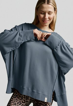 Übergroßer, schieferblauer Pullover mit Rundhalsausschnitt, weiten Ärmeln und rohem Saum, kombiniert mit Leggings im Leopardenmuster.