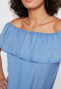 Ljusblå off-shoulder denimtop med volangtröja, som visar en jämn struktur och lätt material. Silverfärgade lagerhalsband synliga.