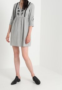 Grå linnklänning med broderad blommönster, v-ringning och trekvartsärmar; kombinerad med svarta träskor. Texturerad tyg, avslappnad passform.