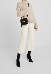 Pull beige en maille, pantalons larges couleur crème, et bottines à bouts pointus noires. Petit sac à main noir avec surface texturée et accents dorés.